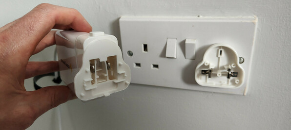 A ChargeASAP Omega 100 watt USB charger, with a UK socket head seen end on. The head has split open. Half is still attached to the charger and half is still plugged into the socket. Live and neutral contacts have been left exposed, connected to 230 volts.