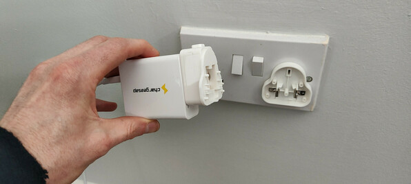 A ChargeASAP Omega 100 watt USB charger, with a UK socket head. The head has split open. Half is still attached to the charger and half is still plugged into the socket. Live and neutral contacts have been left exposed, connected to 230 volts.